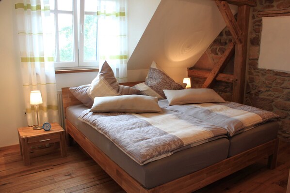 A bedroom with a sloped ceiling, a wooden bed frame, multiple pillows, a bedside table with a lamp, and a window with curtains.