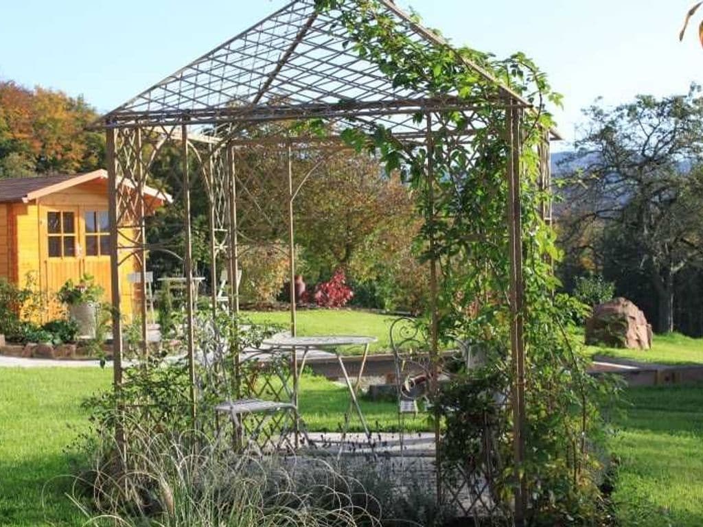 Un jardín con un porche con pérgola, una casa de madera y un elemento de piedra al fondo.