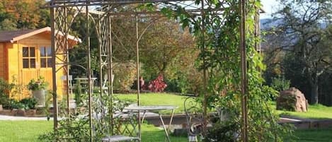 Un jardín con un porche con pérgola, una casa de madera y un elemento de piedra al fondo.
