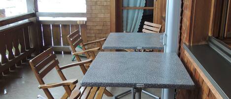 A wooden deck with a table and chairs, a window with curtains, and a view of snow-covered mountains.