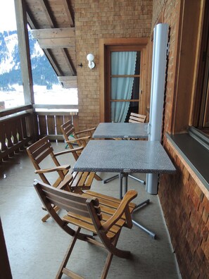 Eine Holzterrasse mit Tisch und Stühlen, ein Fenster mit Vorhängen und Blick auf schneebedeckte Berge.