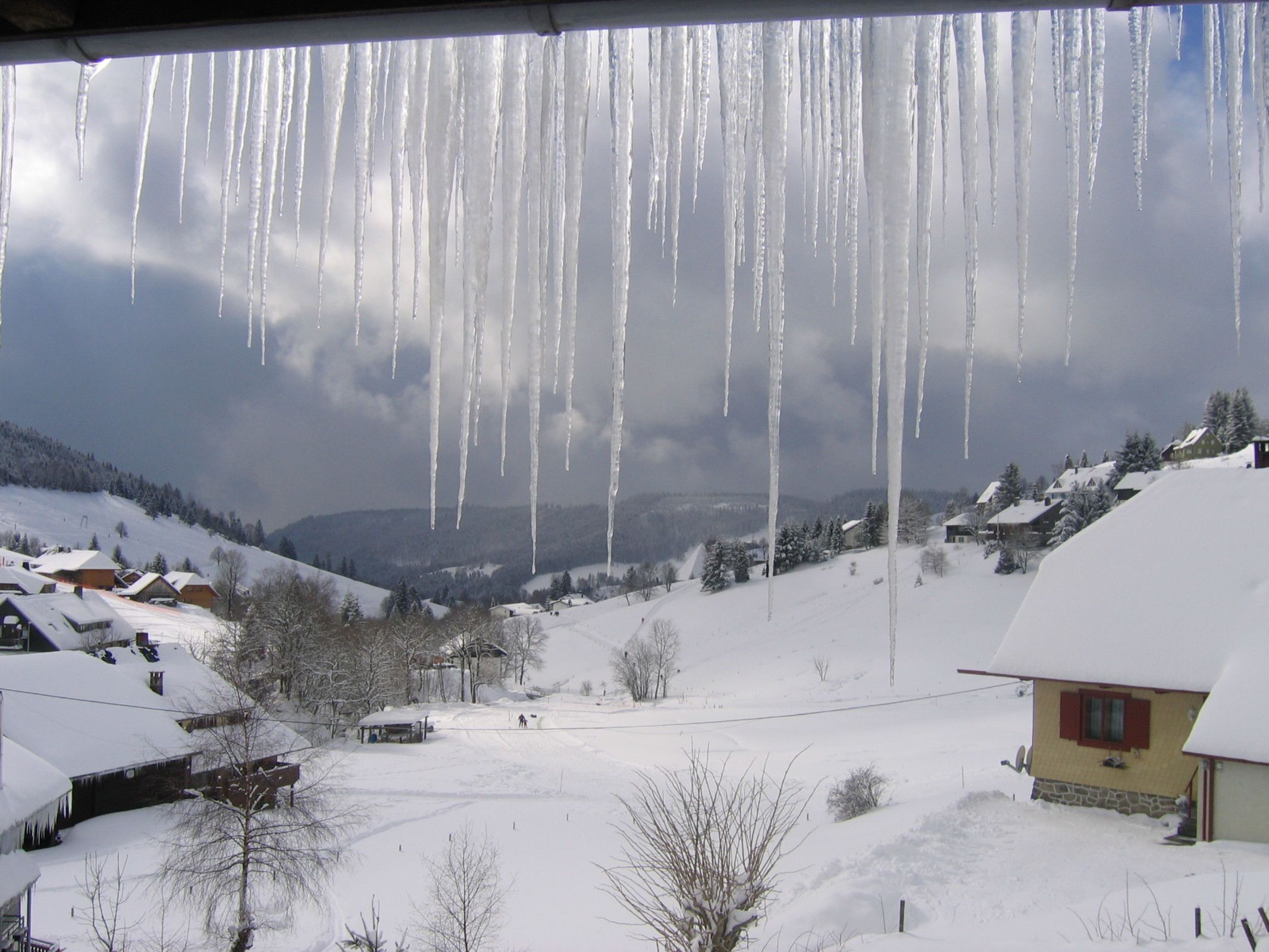 A snowy landscape with icicles hanging from a roof, houses, and a clear sky.