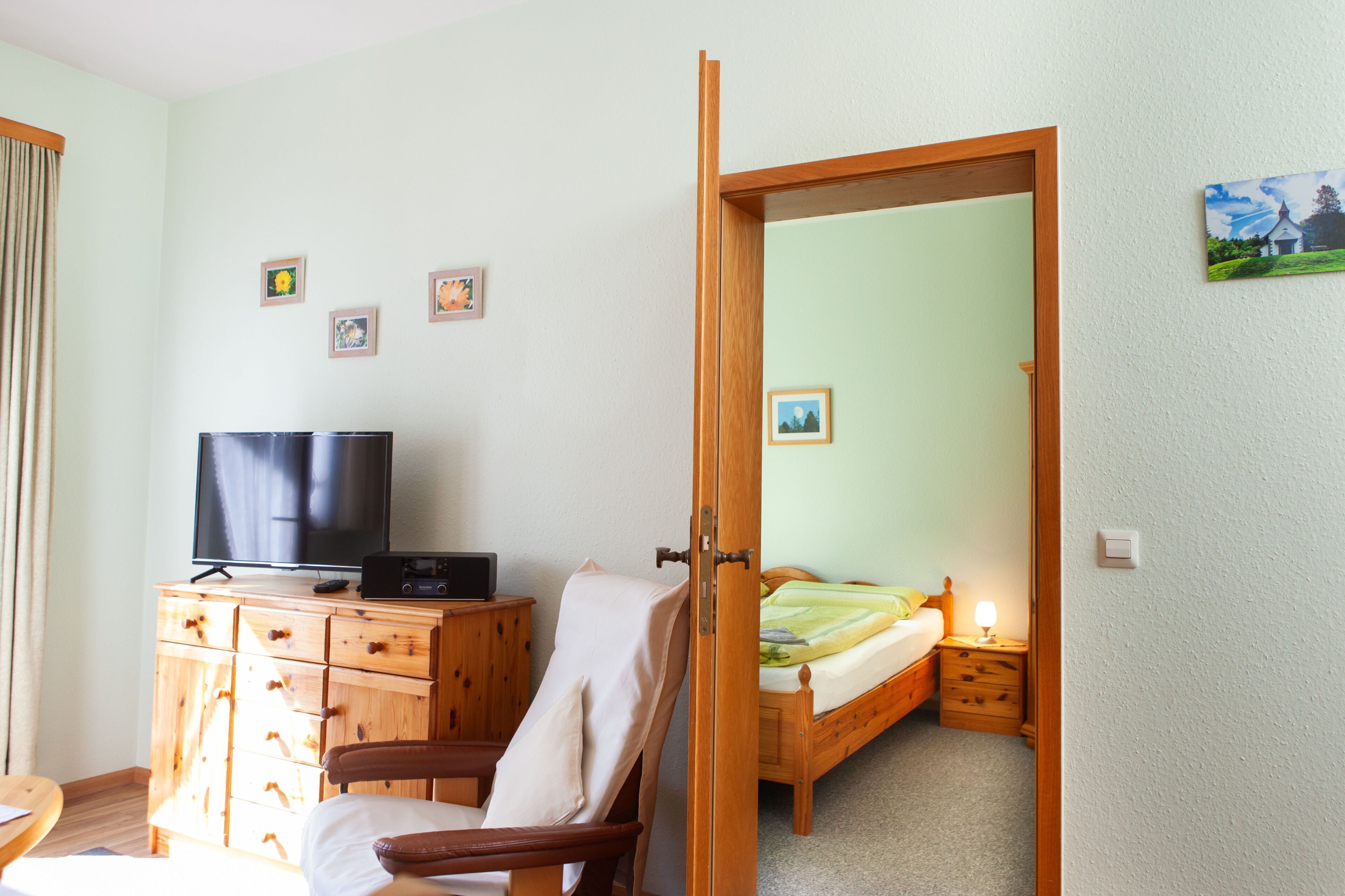 A room with a wooden dresser, a TV, a chair, a bed, and a door leading to another room.