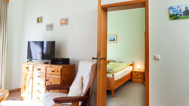 A room with a wooden dresser, a TV, a chair, a bed, and a door leading to another room. - Vacation apartment 3 "Chapel" - Haus Keller vacation apartments (Todtnau)