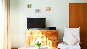 A room with a wooden cabinet, a television, a white chair, and a small table with a plant. - Vacation apartment 3 "Chapel" - Haus Keller vacation apartments (Todtnau)