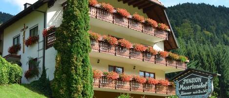 Un immeuble à plusieurs étages, doté de balcons ornés de fleurs, d’une enseigne indiquant qu’il s’agit d’une pension, et d’un jardin luxuriant et verdoyant.