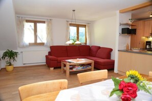 Ein Wohnzimmer mit rotem Sofa, Holztisch und Esstisch mit Stühlen. Ein Fenster mit Vorhängen, eine Pflanze und eine Küchenzeile mit Schränken und einer Mikrowelle.
