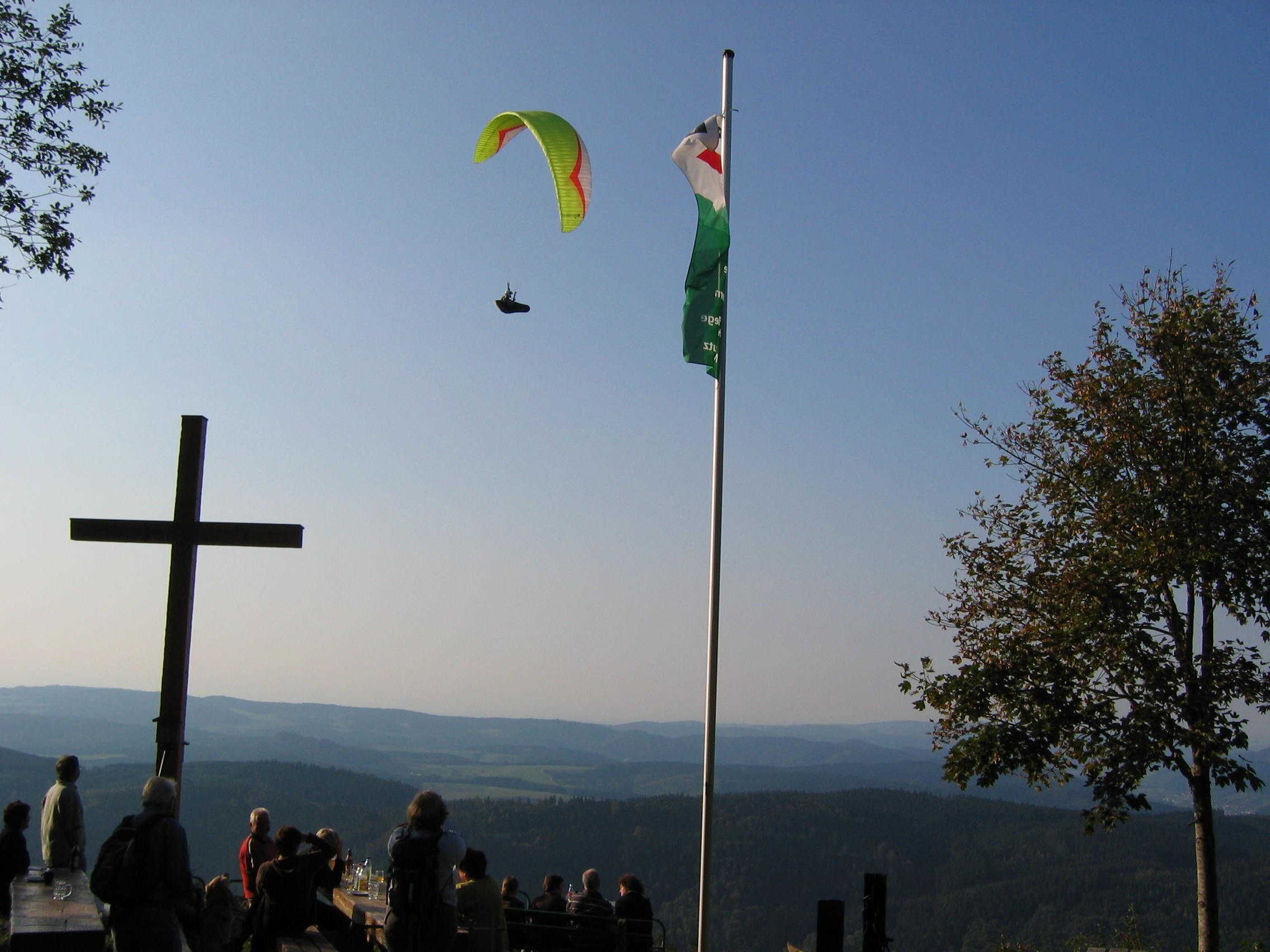 A cross, a flagpole with a flag, and a person paragliding in the background.