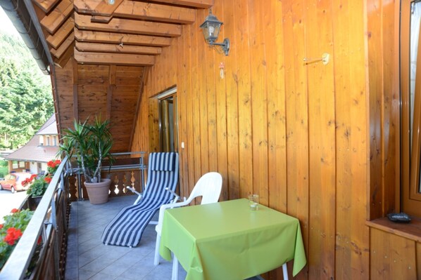Un balcón de madera con una mesa, dos sillas a rayas, una planta en maceta y una puerta.