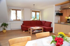 Ein Wohnzimmer mit rotem Sofa, Holztisch und Esstisch mit Stühlen. Ein Fenster mit Vorhängen, eine Pflanze und eine Küchenzeile mit Schränken und einer Mikrowelle.