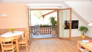 Una habitación con una mesa de madera, sillas, un sofá, un ventanal corredero que da a un área exterior con terraza de madera, un televisor y una planta en maceta.