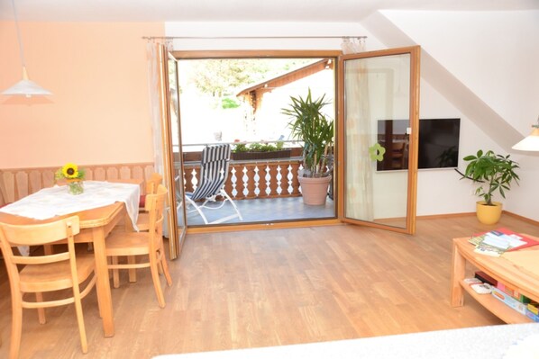 Ein Zimmer mit Holztisch, Stühlen, einer Couch, einer Glastür, die zu einem Außenbereich mit Holzterrasse führt, einem Fernseher und einem Blumentopf.