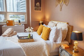 A hotel room with a bed, a bedside table with a lamp, a vase with flowers, and a view of the city through the window.