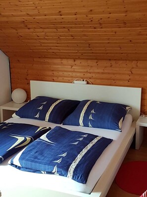 Un lit avec une tête de lit blanche, une literie bleue ornée de motifs de voiliers et un tapis rouge sur le sol.