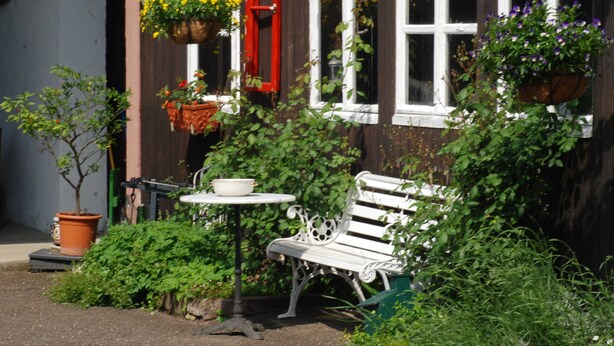Un banco blanco y una mesa frente a un edificio con contraventanas rojas y cestas de flores colgantes.