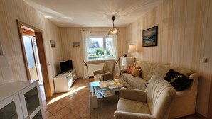 Ein Wohnzimmer mit Sofa, Sesseln, Couchtisch, Fernseher und einem Fenster mit Vorhängen.