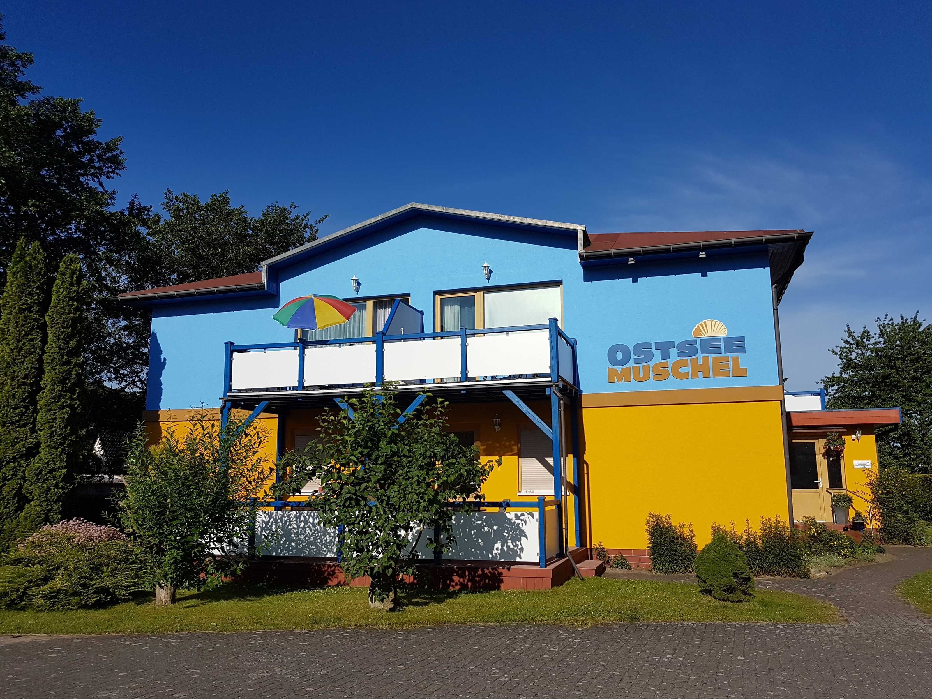 A building with a blue and yellow facade, a sign reading 'OSTSEE MUSCHEN,' and a balcony with a colorful umbrella.