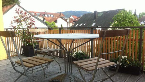 Una terraza de madera con una mesa y sillas de metal, rodeada por una valla de madera y plantas en macetas.