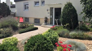 Een modern gebouw met een plat dak, een overdekte ingang en een kleine tuin met diverse planten en bloemen.