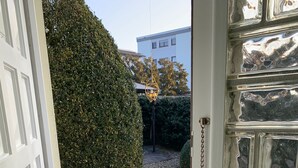 Een deur met een metalen hekwerk, een witte deurpost, en een kleine tuin met een haag, een plant in pot en een metalen bank.