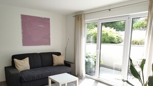A modern living room with a dark grey sofa, a small white table, and a sliding glass door leading to an outdoor area with plants.