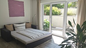 A modern bedroom with a large bed, a sliding glass door to the balcony, and a potted plant.