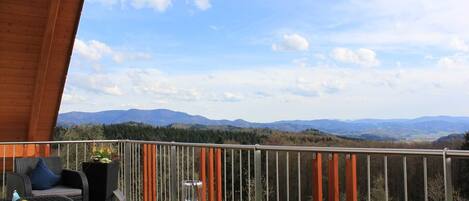 A balcony with wicker furniture, a glass table, and a view of distant mountains.