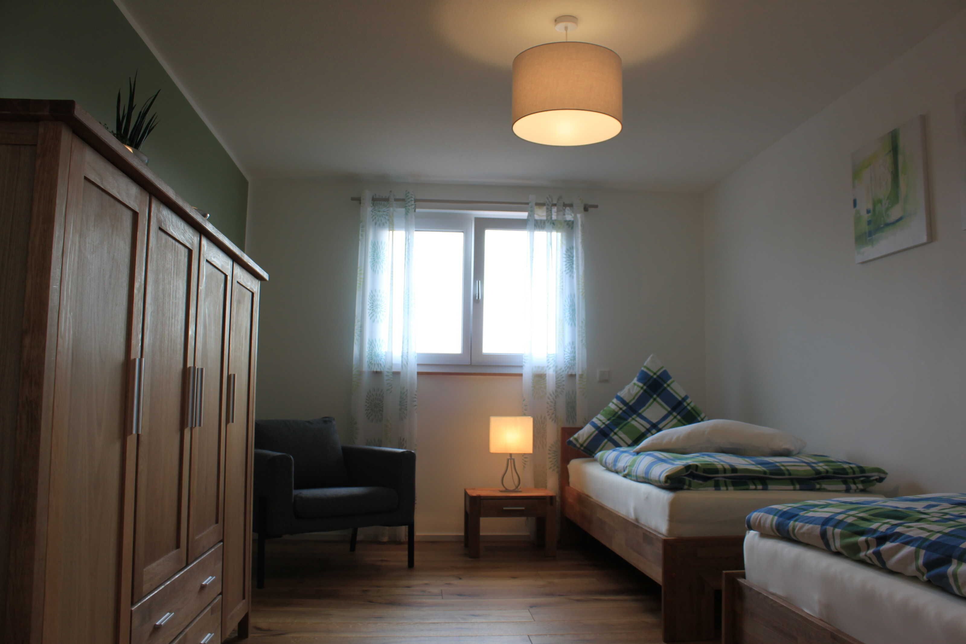 Una habitación con un armario de madera, una mesita con una lámpara, un sofá con ropa de cama a cuadros y una ventana con cortinas translúcidas.