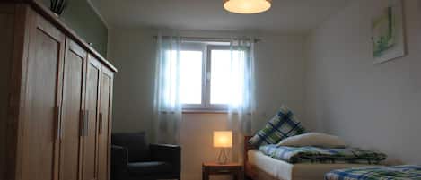 Una habitación con un armario de madera, una mesita con una lámpara, un sofá con ropa de cama a cuadros y una ventana con cortinas translúcidas.