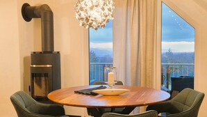 A modern dining area with a round wooden table, two upholstered chairs, a black wood-burning stove, and a large window with a view of mountains. - flammende.aussicht, Germany (Freiamt)