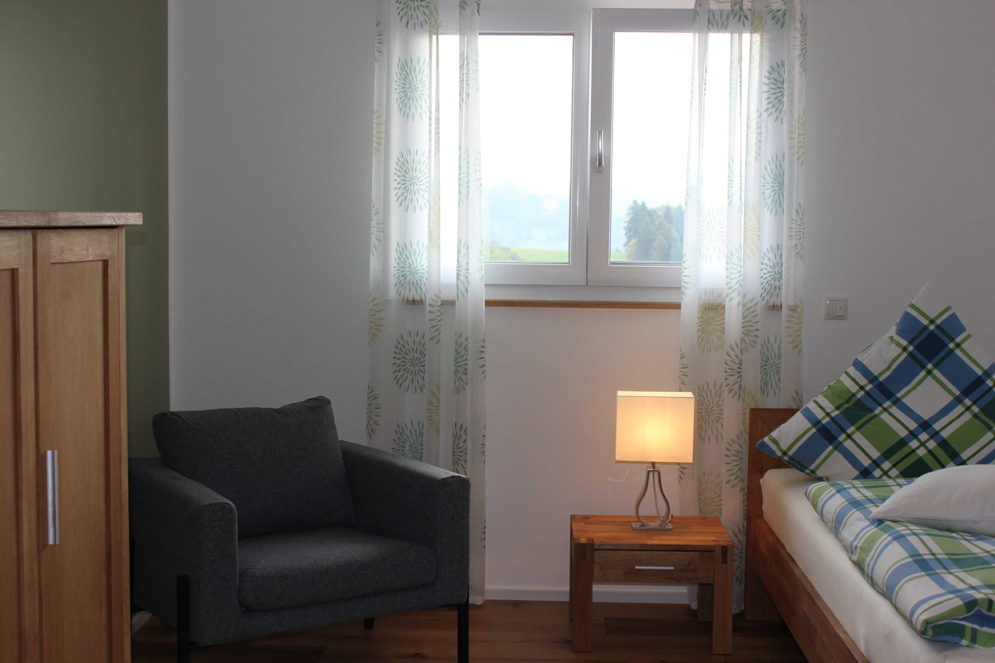 Ein Zimmer mit einer Couch, einem Nachttisch aus Holz mit Lampe, einem Bett mit Karo-Bettwäsche und einem Fenster mit Vorhängen.