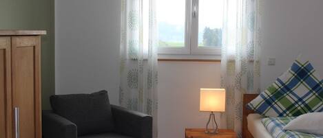 Ein Zimmer mit einer Couch, einem Nachttisch aus Holz mit Lampe, einem Bett mit Karo-Bettwäsche und einem Fenster mit Vorhängen.