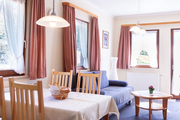 A room with a dining table, chairs, a sofa, a small table with a plant, and a window with curtains.