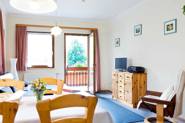 A room with a dining table, chairs, a TV, a wooden cabinet, and a view of a balcony with a tree.