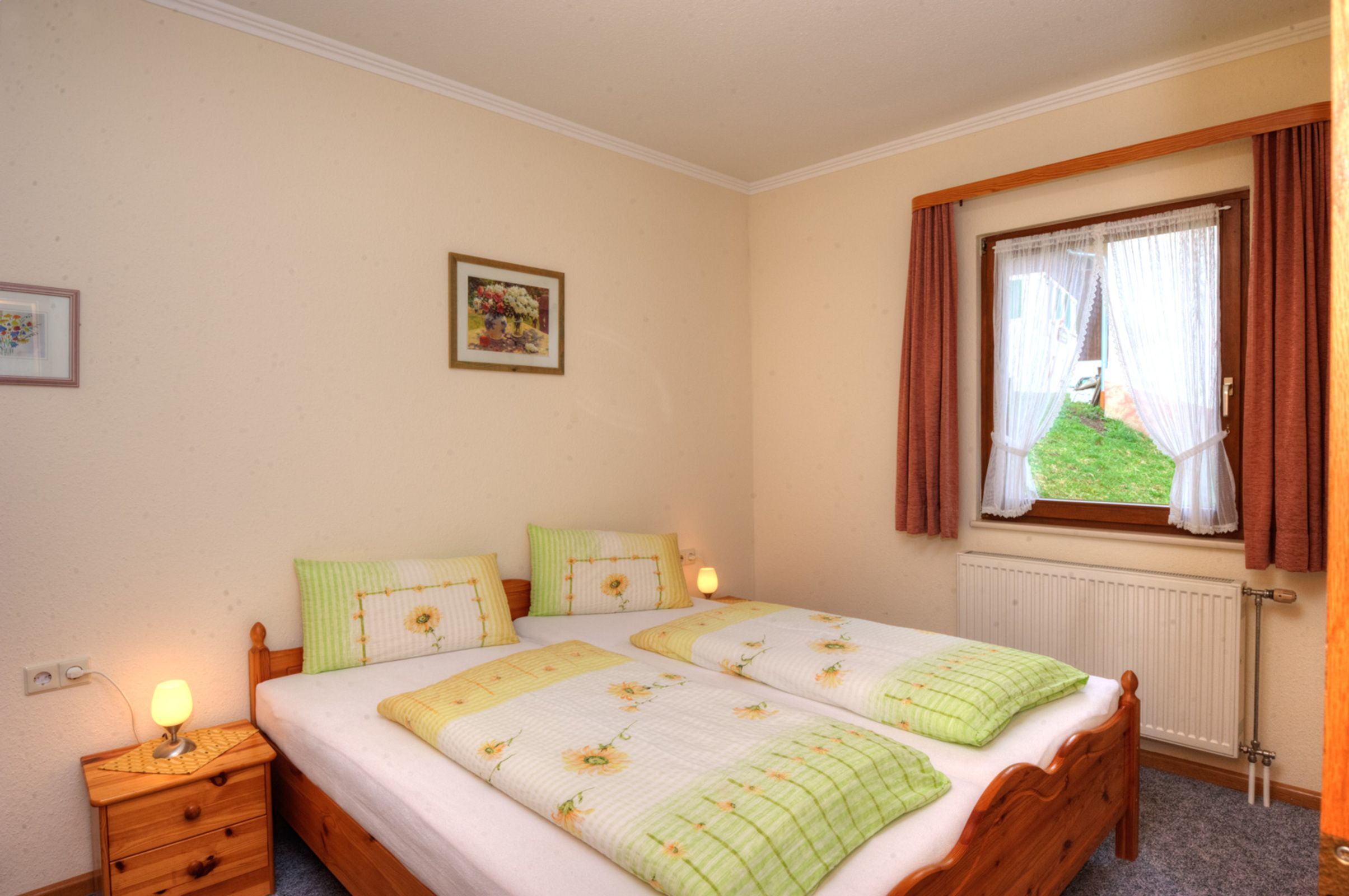 A bedroom with two beds, a nightstand, a window with curtains, and a radiator.
