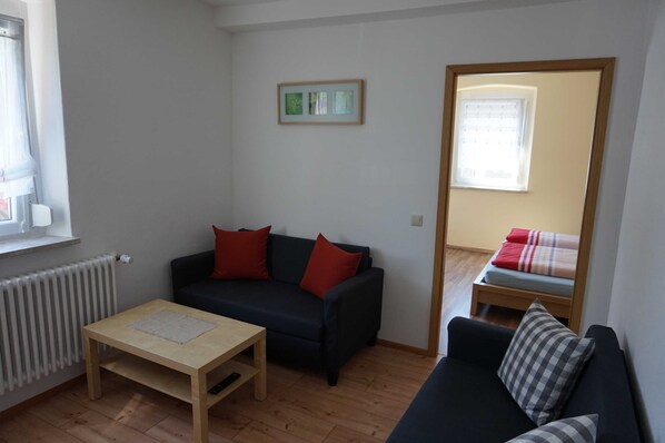 A small, well-lit room with a dark-colored sofa, a wooden table, and a window with a view of a bed.