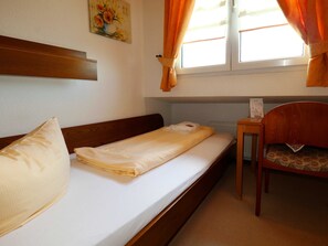 Un llit individual amb capçalera de fusta, una taula petita amb una cadira, i una finestra amb cortines.