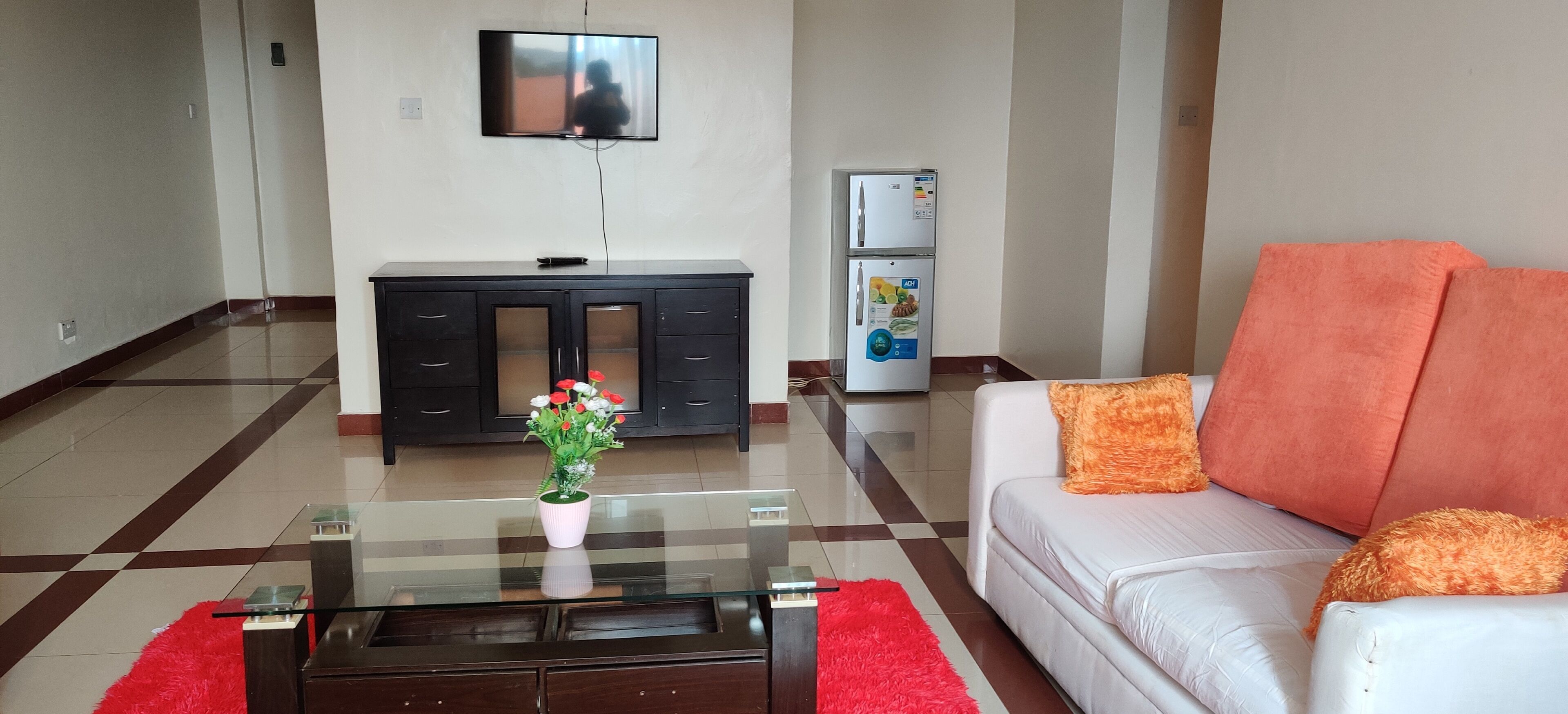 A modern living room with a glass coffee table, white sofa, and orange cushions. A black entertainment unit with a TV and a refrigerator is in the background.