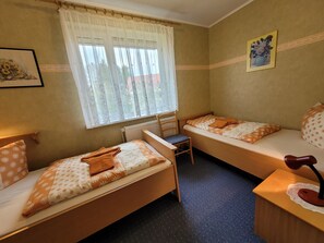 A room with two single beds, a chair, a desk with a lamp, a radiator, and a window with sheer curtains.