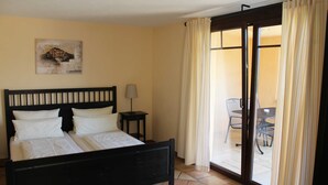 A bedroom with a bed, a nightstand, a lamp, and a sliding glass door leading to a balcony with a table and chairs. - Two-Bedroom Apartment (5) - Pension Vanii (Rust)
