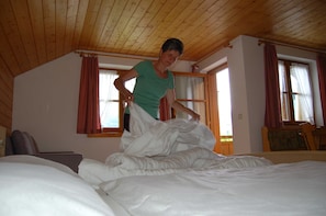 A room with a wooden ceiling, a bed with white bedding, a chair, and a window with curtains.
