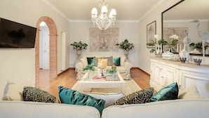 A living room with a sofa, coffee table, and decorative items. There is a television mounted on the wall and a chandelier hanging from the ceiling. - Spacious Home with Elevator along Via Guinigi inside the Lucca Walls (Lucca)