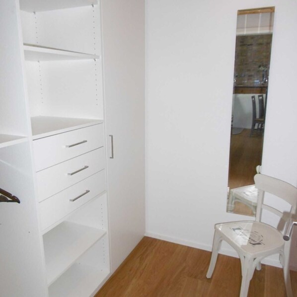 A white closet with open shelves and drawers, a wooden floor, and a white chair in a corner. - Apartment 3 - Guesthouse Andrea Herzig (Breisach am Rhein)