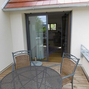 A balcony with a round metal table and two wicker chairs, a sliding glass door leading to a kitchen, and a wooden deck. - Apartment 3 - Guesthouse Andrea Herzig (Breisach am Rhein)