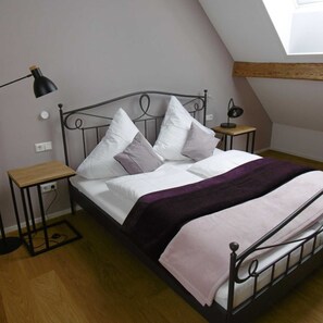 A single bed with a metal frame, white bedding, and multiple pillows. There are two bedside tables, one with a lamp on it. The room has wooden flooring and a sloped ceiling with exposed beams. - Apartment 3 - Guesthouse Andrea Herzig (Breisach am Rhein)