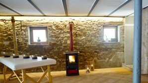 Ein Zimmer mit Steinwänden, einem Kaminofen, einem Tisch mit Schalen und zwei Fenstern.