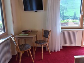 A room with a TV mounted on the wall, a table with chairs, a radiator, and a window with curtains. - Double room 3 with toilet and shower, 1 - 3 persons - Mosertonihof (Elzach)
