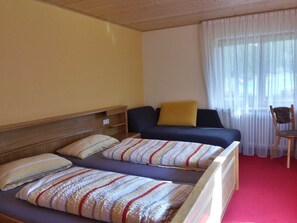 Habitación de hotel con dos camas, cabecero de madera, sofá con un cojín amarillo, una silla y una ventana con cortinas traslúcidas.