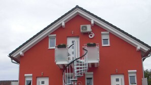 Un bâtiment rouge avec un balcon blanc, un escalier en colimaçon et une petite fenêtre ornée d’une plante.