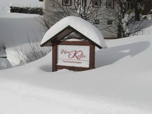 Un panneau recouvert de neige indiquant « Hans Keller Ferienwohnungen », avec une structure en bois et un toit également recouvert de neige.